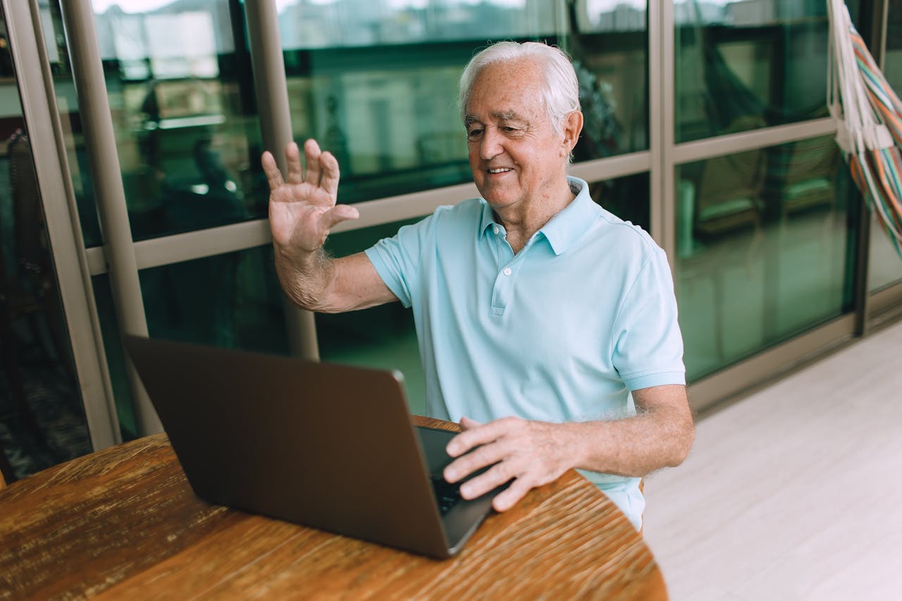 Elderly man happily waving during a video call on his laptop, seated at home.