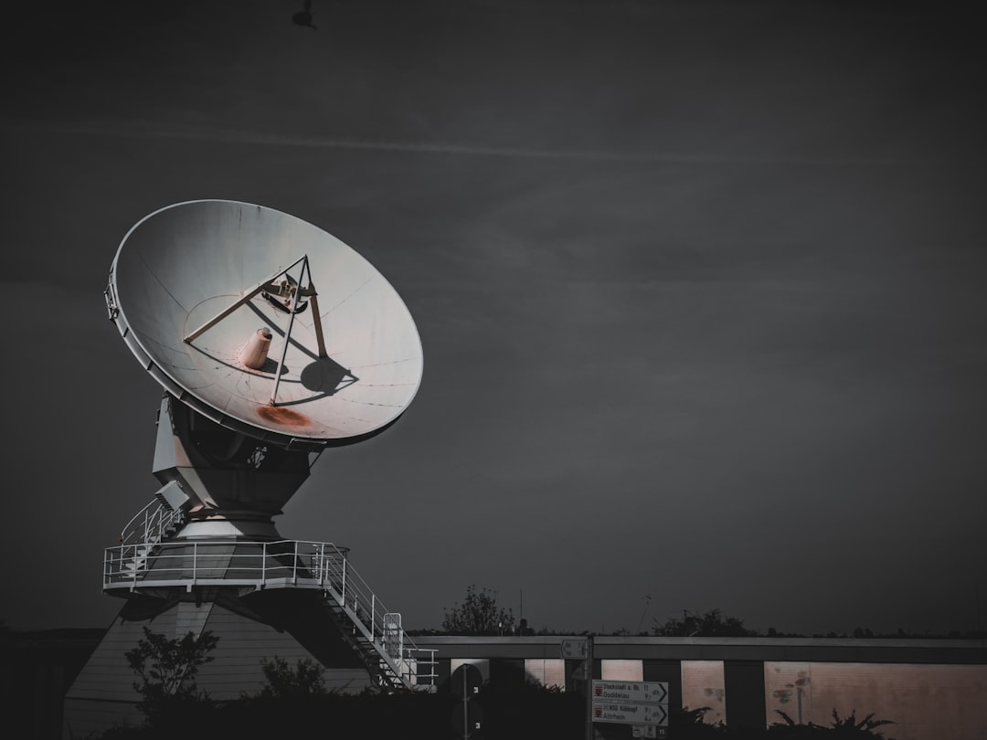 An imposing satellite dish is photographed against a dusky sky, featuring a prominent steel framework and the dishs parabolic shape designed for signal reception and transmission. The scene is composed with the satellite dish dominating the upper half of the frame, underscored by a building with directional signage partially visible below.